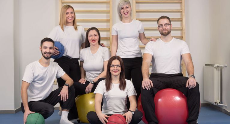 Gruppenbild Therapeutinnen und Therapeuten in weißen T-Shirts mit Bällen vor einer Sprossenwand