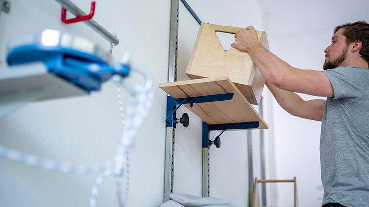 Patient stellt eine Kiste auf ein Regal in der medizinisch-beruflich orientierten Rehabilitation