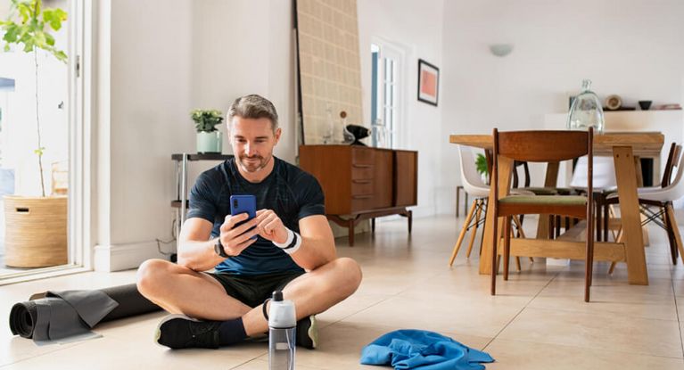 Mann in Sportkleidung prüft nach dem Indoor-Training im Wohnzimmer die Ergebinsse auf seinem Smartphone