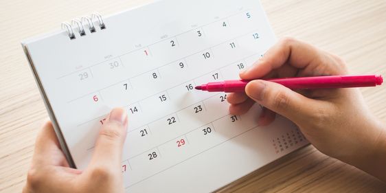 Eine Person markiert mit einem pinkfarbenen Stift ein Datum in einem Tischkalender. Der Kalender zeigt einen Monatsüberblick mit Zahlen von 1 bis 31, wobei die Sonntage in rot markiert sind. Die Person hält den Kalender mit einer Hand fest, während sie mit der anderen Hand den Stift führt.